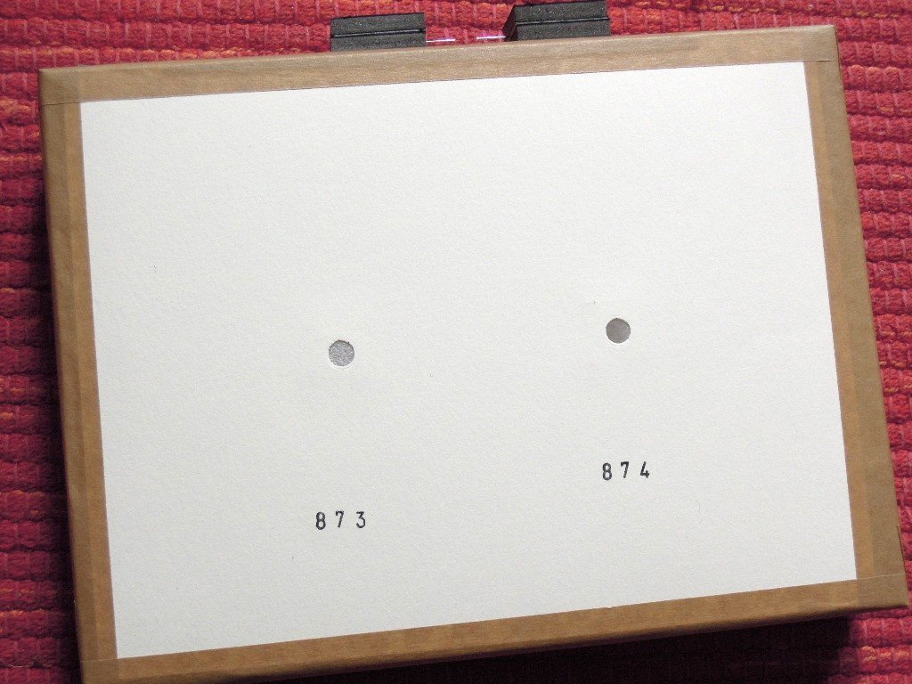 White cardboard camera with brown paper tape on red cloth, two pierced holes side by side, the stamped numbers 873 and 874 below them.