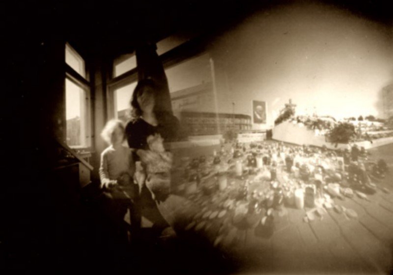Sepia pinhole photograph made from two exposures on one negative: on the left a woman with a small child at a window, on the right a wide square densely covered in grave candles, with a portrait on a wall behind it.
