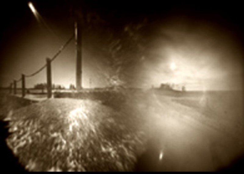 Sepia pinhole image made of two exposures: a fence and a telegraph pole against a low sun on the left, a glaring bright plain on the right, the two halves merging into one another.