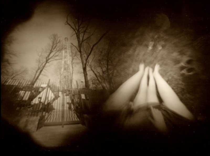 Sepia pinhole photograph from two exposures: on the left a wrought-iron gate between bare trees with a lattice tower behind it, on the right the brightly lit, raised legs of a reclining person.