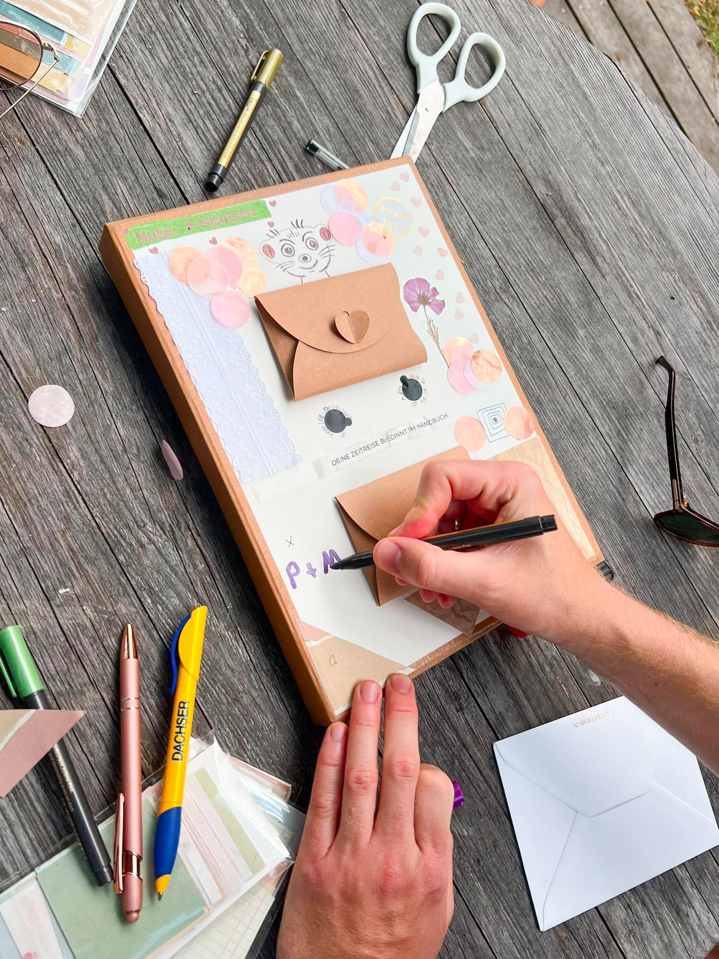 Hands decorating a Both In Time camera at a wooden table with paper scraps, envelopes and pens.