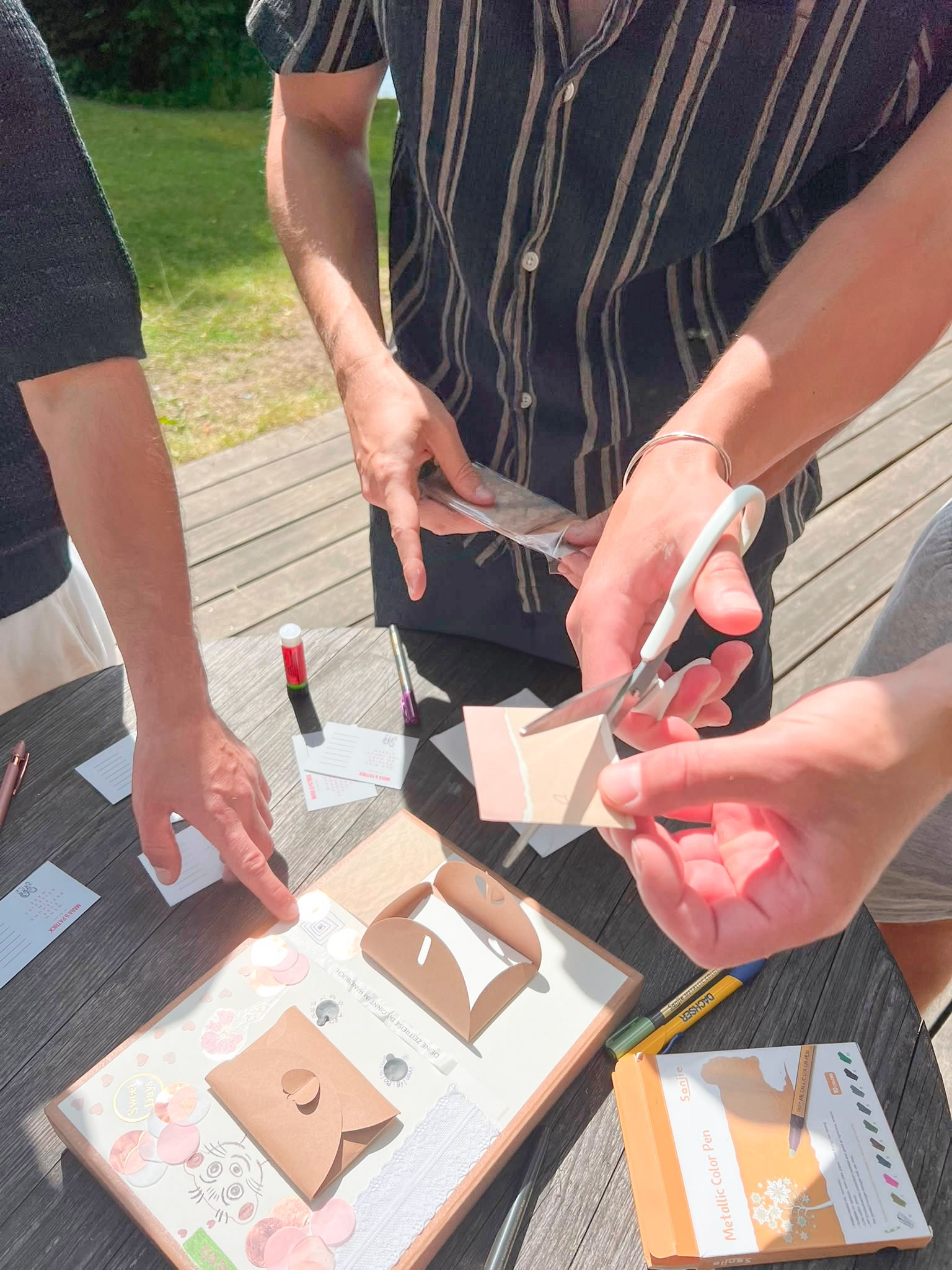 Zwei Personen bauen an einem Holztisch gemeinsam an einer Both In Time Kamera, zwischen Schere, Stiften und Papier.