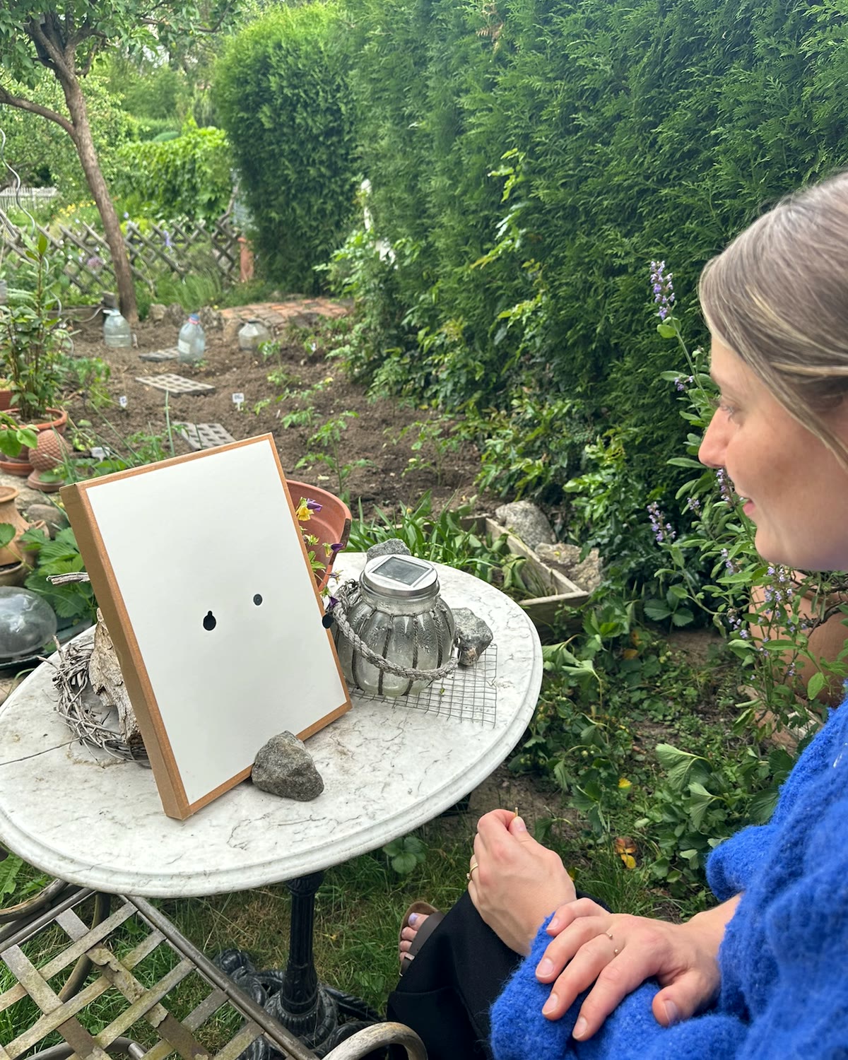 Eine Frau im blauen Pullover sitzt an einem steinernen Gartentisch, vor ihr steht aufrecht die handgefertigte Both In Time Lochkamera aus Karton mit zwei sichtbaren Öffnungen.