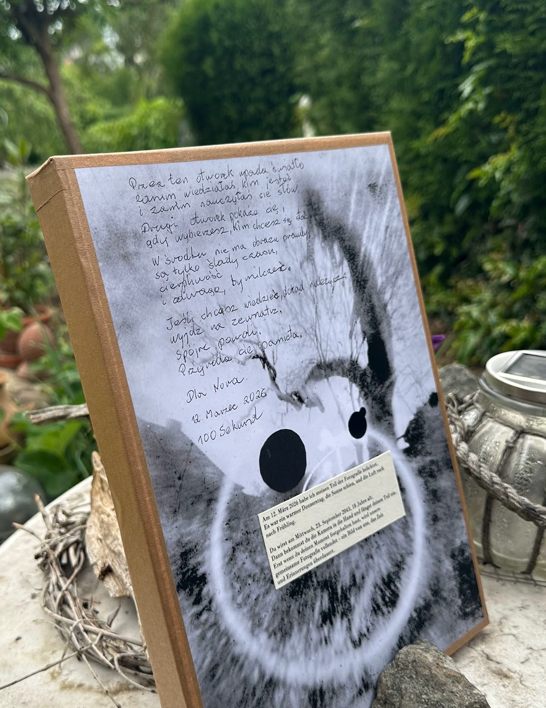 Una cámara Both In Time sobre una mesa de piedra al aire libre: la primera mitad ya está expuesta, con una dedicatoria manuscrita con fecha y una nota pegada que promete la segunda exposición para 2043.