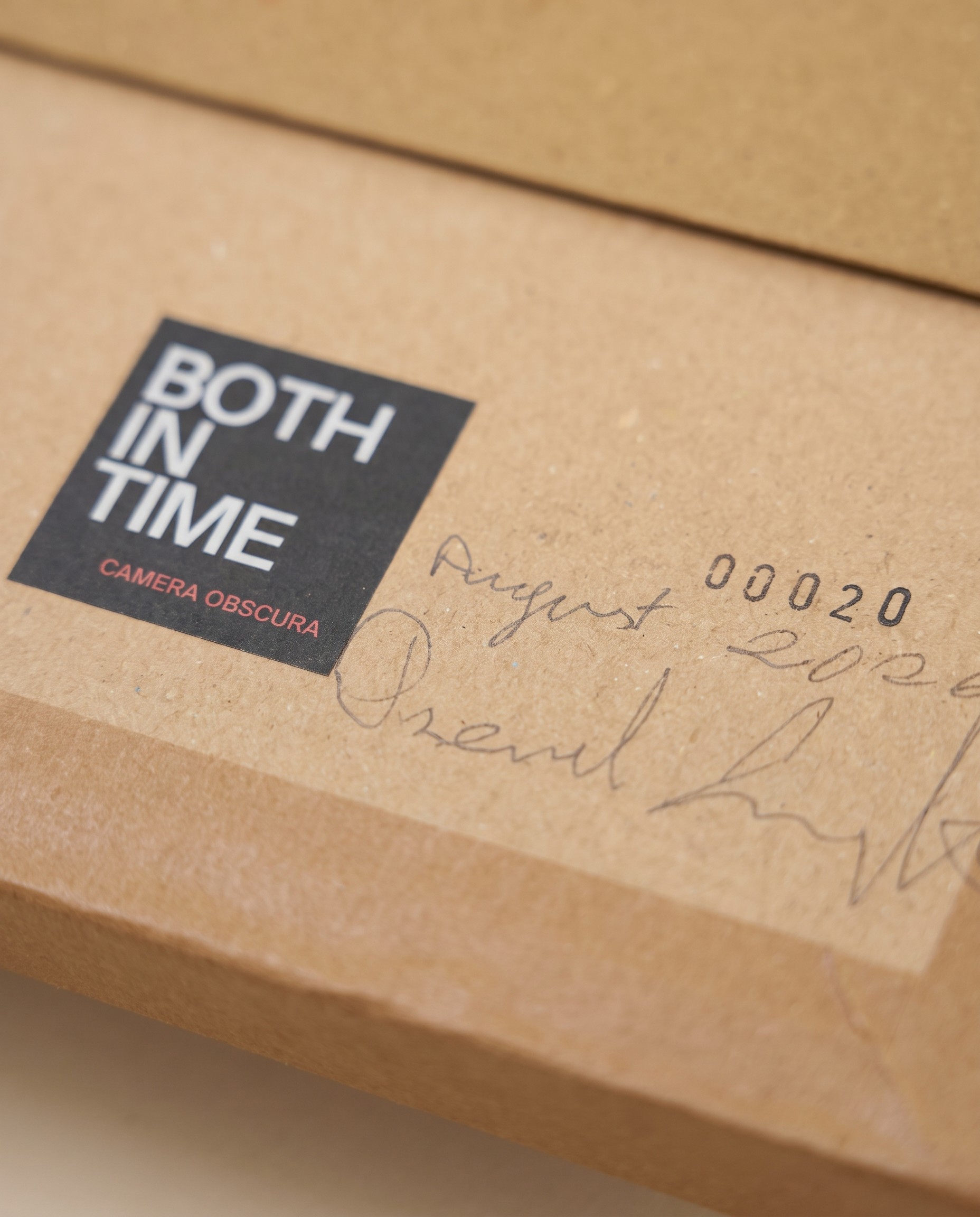 Nahaufnahme einer Both In Time Kamera aus Karton: schwarzes Logo-Etikett, gestempelte Nummer 00020, daneben ein handschriftliches Datum und eine Unterschrift in Bleistift.